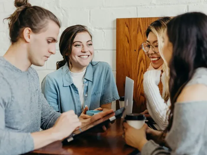 Group of young professionals smiling and discussing ideas over coffee at wooden table with laptop and tablet